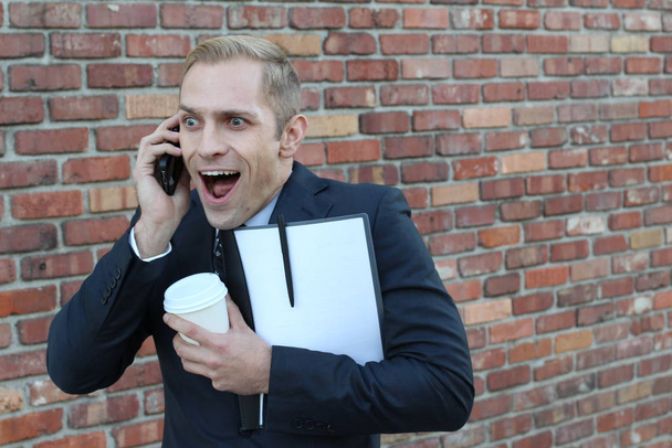 close-up shot of handsome adult businessman with folder and paper cup of coffee talking by phone in front of brick wall on street - Photo, Image