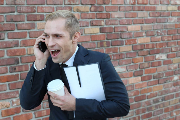 close-up shot of handsome adult businessman with folder and paper cup of coffee talking by phone in front of brick wall on street - Φωτογραφία, εικόνα