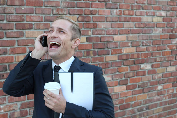 primer plano de hombre de negocios adulto guapo con carpeta y taza de papel de café hablando por teléfono en frente de la pared de ladrillo en la calle
 - Foto, Imagen