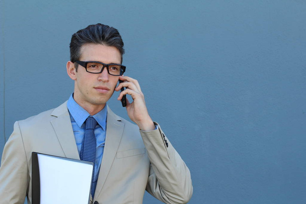 close-up portrait of handsome young businessman in beige suit talking by phone in front of blue wall - Fotó, kép