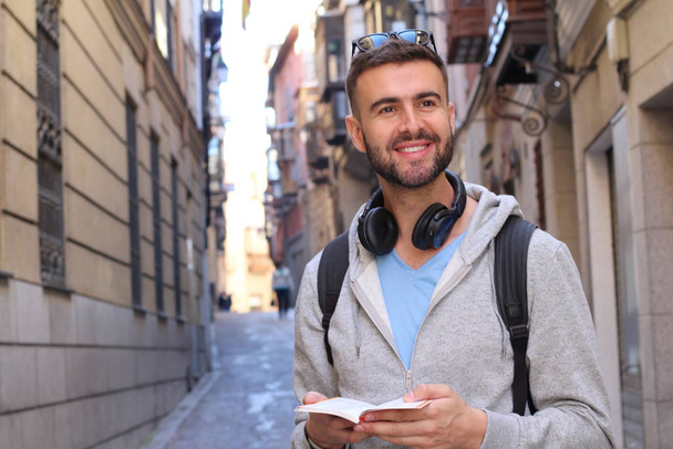 close-up portrait of handsome young man with backpack and book on city street - Фото, изображение