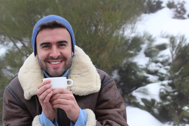 retrato cercano de un joven guapo vestido con ropa caliente con una taza de bebida caliente en la naturaleza en invierno. - Foto, Imagen