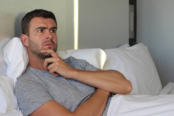 close-up portret van knappe jonge expressieve man aanraken van zijn kin in bed thuis - Foto, afbeelding