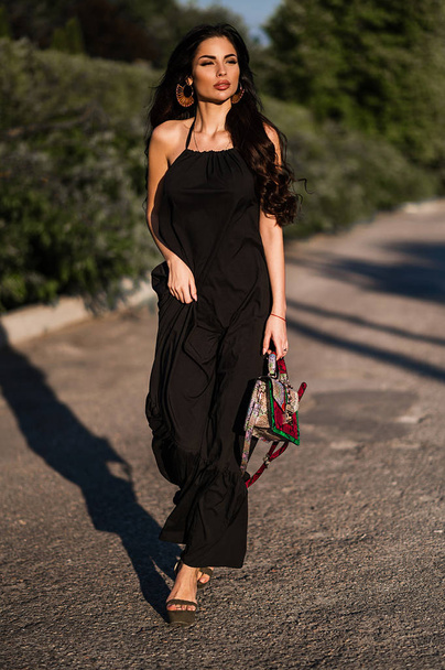 A brunette in a long black dress is walking along the streets of the city. Portrait of a brunette in a lush afternoon in a dress - Foto, imagen