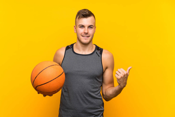 Jeune homme blond beau tenant une boule de panier sur fond jaune isolé pointant vers le côté pour présenter un produit
 - Photo, image