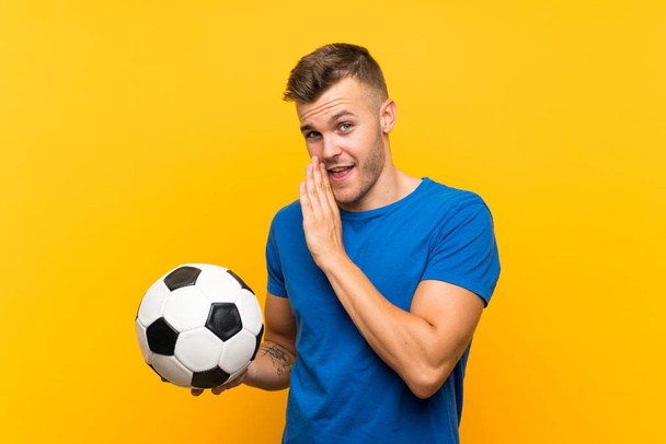 Jeune homme blond beau tenant un ballon de football sur fond jaune isolé chuchotant quelque chose
 - Photo, image