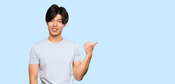 Hombre asiático con camisa azul apuntando hacia un lado para presentar un producto sobre fondo azul aislado
 - Foto, Imagen