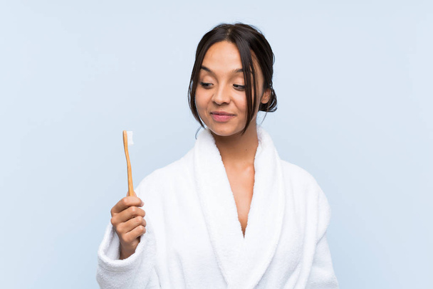 Young brunette woman in bathrobe brushing her teeth over isolated blue background with happy expression - Foto, Imagem