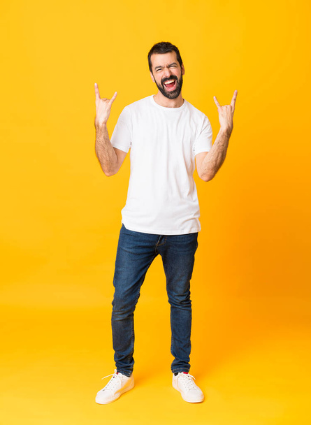 Ganzkörperaufnahme eines Mannes mit Bart vor isoliertem gelben Hintergrund, der eine Rock-Geste macht - Foto, Bild
