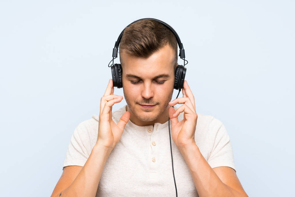 junger schöner blonder Mann über isoliertem blauem Hintergrund, der mit Kopfhörern Musik hört - Foto, Bild