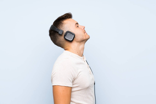Νέος όμορφος ξανθός άνθρωπος πάνω από απομονωμένο μπλε φόντο ακούγοντας μουσική με ακουστικά - Φωτογραφία, εικόνα