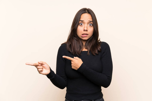 Young brunette woman with white sweater over isolated background frightened and pointing to the side - Photo, Image