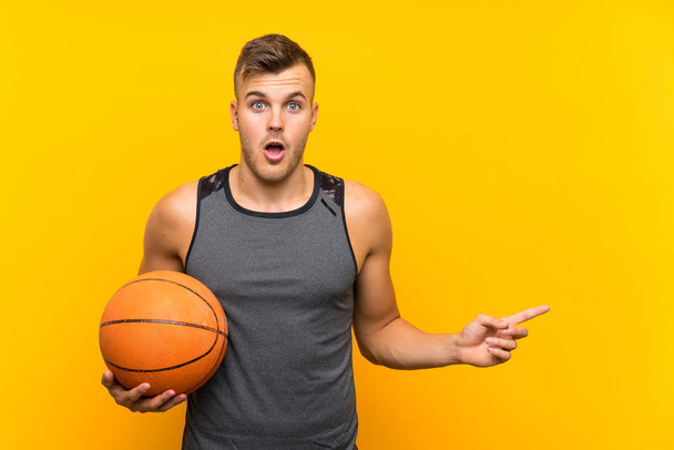 Young handsome blonde man holding a basket ball over isolated yellow background surprised and pointing side - Фото, изображение