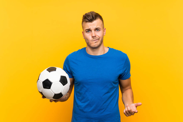 Giovane bell'uomo biondo che tiene un pallone da calcio su uno sfondo giallo isolato facendo dubbi gesto mentre solleva le spalle
 - Foto, immagini