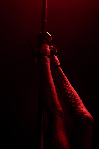 Ausgeschnittene Ansicht von Stripperin in Heels in der Nähe von Pylon auf schwarz mit roter Beleuchtung  - Foto, Bild