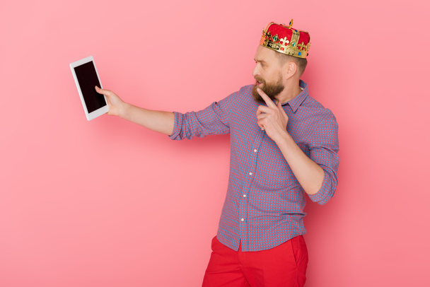 pensive man with crown looking at digital tablet on pink background  - Photo, Image
