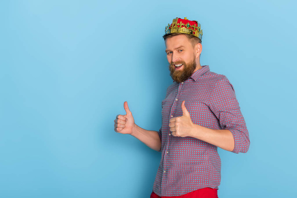 smiling man with crown showing thumbs up on blue background  - Photo, Image