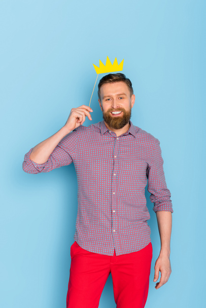 usmívající se a pohledný muž drží papírovou korunu na modrém pozadí  - Fotografie, Obrázek