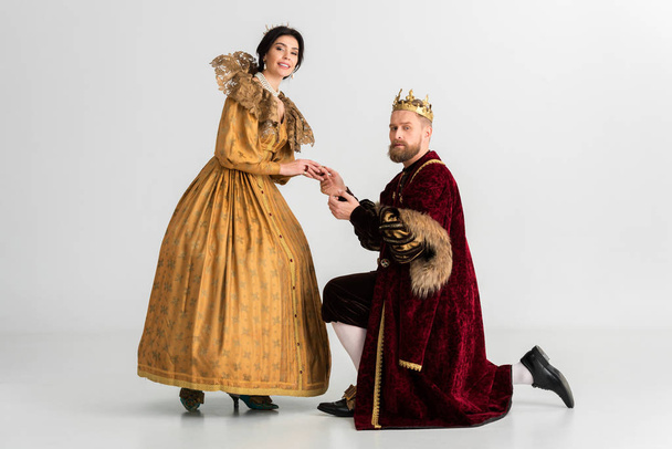 king with crown bending on knee and holding hand of smiling queen on grey background  - Photo, Image