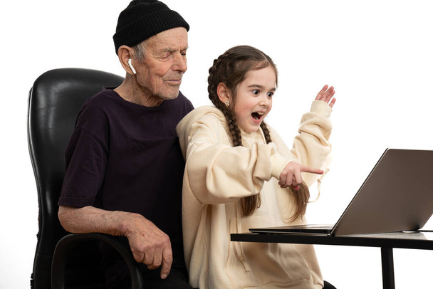 glückliche Enkelin in beigem Kapuzenpulli spielt mit ihrem stilvollen Großvater in schwarzem Hut und T-Shirt am Laptop, verbringt Zeit miteinander, isoliert vor weißem Hintergrund - Foto, Bild