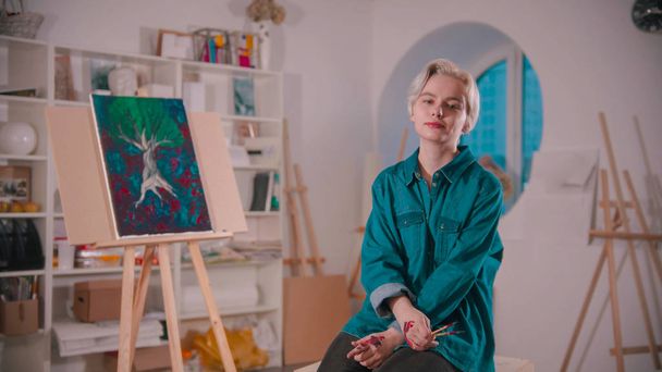Μια νεαρή γυναίκα καλλιτέχνης με κοντά ξανθά μαλλιά κάθεται στο στούντιο τέχνης - Φωτογραφία, εικόνα