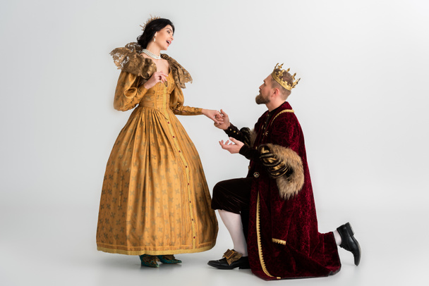 king with crown bending on knee and holding hand of shocked queen on grey background  - 写真・画像