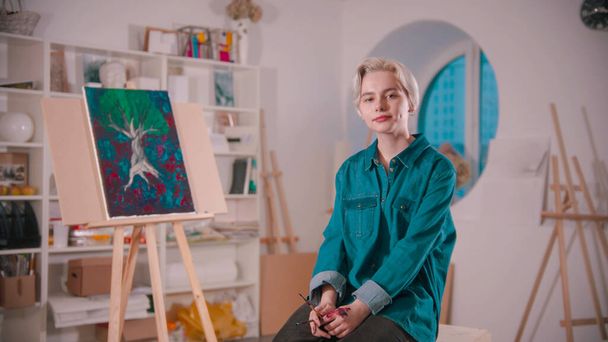 Uma jovem artista com cabelo loiro curto sentada no estúdio de arte sua pintura no fundo
 - Foto, Imagem