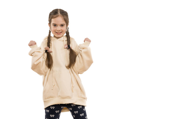 Ευτυχισμένο νεαρό κορίτσι με κοτσιδάκια ντυμένο με μπεζ κουκούλα, σε λευκά ασύρματα ακουστικά, ενθουσιασμένοι κάνει χειρονομία νικητής με τα χέρια ψηλά, χαμογελώντας και ουρλιάζοντας για την επιτυχία. Έννοια εορτασμού. - Φωτογραφία, εικόνα