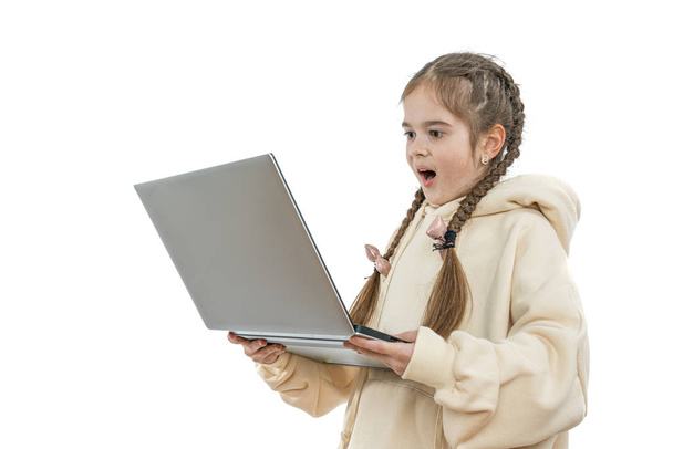 Σοκαρισμένη νεαρή μαθήτρια με κοτσίδες, ντυμένη με μπεζ κουκούλα κρατά το λάπτοπ στα χέρια και το κοιτάζει, απομονωμένη σε λευκό φόντο - Φωτογραφία, εικόνα