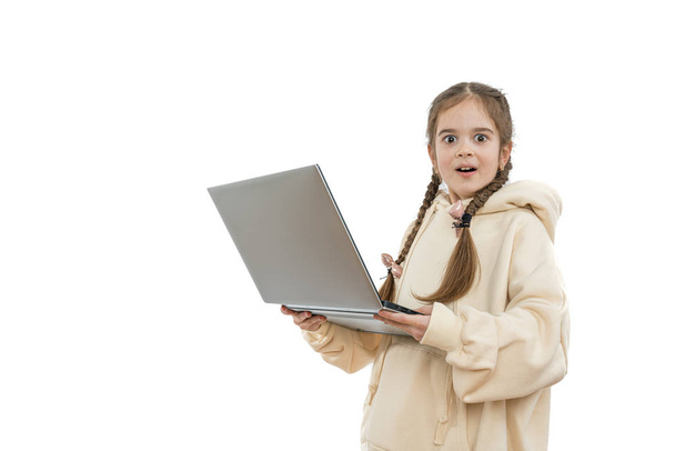 Lenyűgözött és izgatott fiatal lány diák copfos, öltözött bézs kapucnis tartja laptop a kezében, és nézi a kamera, elszigetelt fehér háttér - Fotó, kép