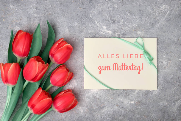 "Alles Liebe zum Muttertag", that means in German "All the love on Mother's day". Text on paper card. Flat lay with red tulips, green decorative flowers and paper card on grey textured background. - Фото, изображение