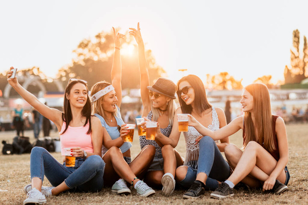 smiling girlfriends with drinks sitting on grass on music festival  - Photo, Image