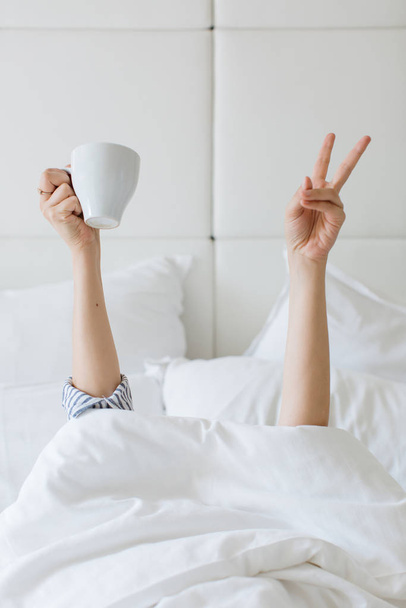 person holding white cup and raising two fingers up in white bed - 写真・画像