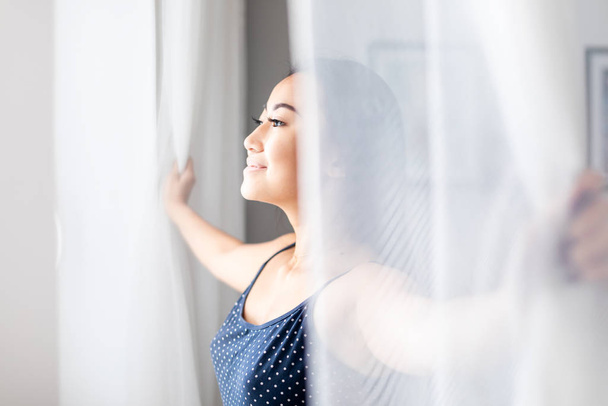 Азійська дівчинка в піжамі відчиняє віконні фіранки вдома вранці. - Фото, зображення