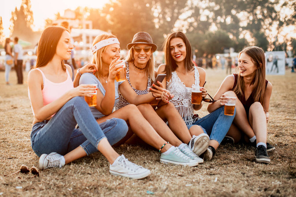 smiling girlfriends with drinks sitting on grass on music festival  - Zdjęcie, obraz