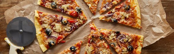 вид сверху на срезанную вкусную итальянскую пиццу с оливками на выпечке бумаги рядом с ножом для пиццы на деревянном фоне, панорамный снимок
 - Фото, изображение