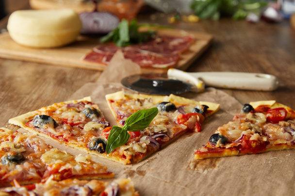 selective focus of cut tasty italian pizza with olives on baking paper near ingredients and pizza knife on wooden table - Foto, afbeelding