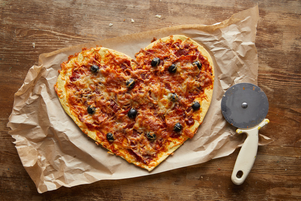 top view of delicious italian pizza in heart shape on baking paper near pizza knife on wooden table - Fotoğraf, Görsel
