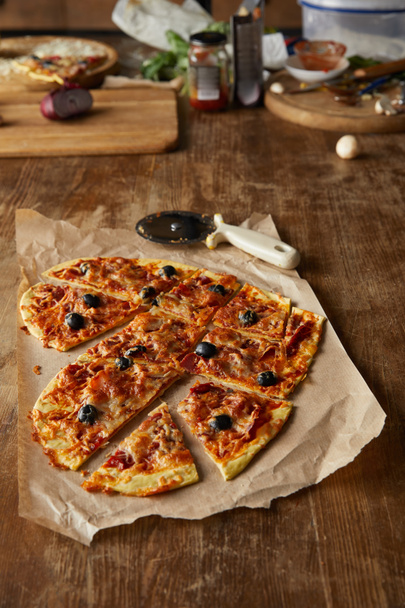 leckere italienische Pizza in Herzform in Stücke geschnitten auf Backpapier neben Pizzamesser auf Holztisch - Foto, Bild