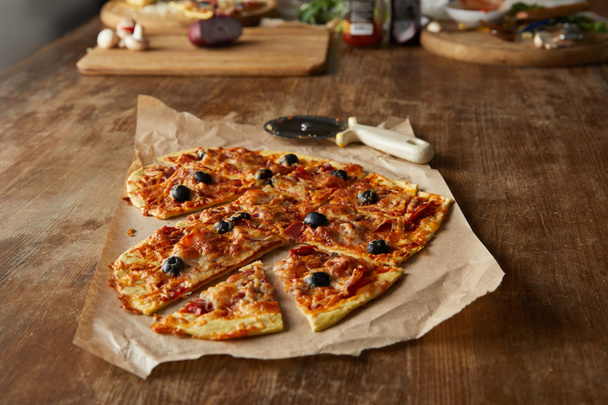 délicieuse pizza italienne en forme de coeur coupée en morceaux sur du papier cuisson près d'un couteau à pizza et des ingrédients sur une table en bois
 - Photo, image