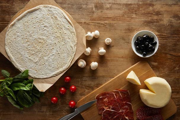 сверху вид на сырое тесто, помидоры черри, базилик, грибы, оливки, моцарелла, салями и прошутто для пиццы на деревянном столе
 - Фото, изображение