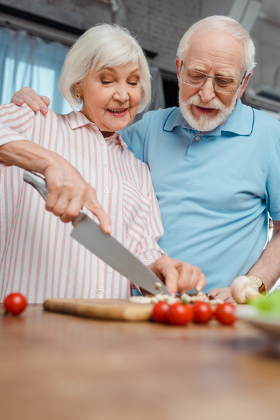 Selective focus of senior man hugging wife during cooking on kitchen table - Fotó, kép
