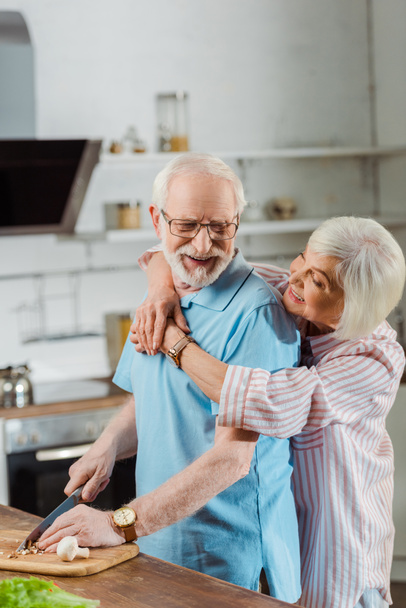 キッチンテーブルの上で野菜を切りながら夫を抱きしめる笑顔の女性の側面図 - 写真・画像