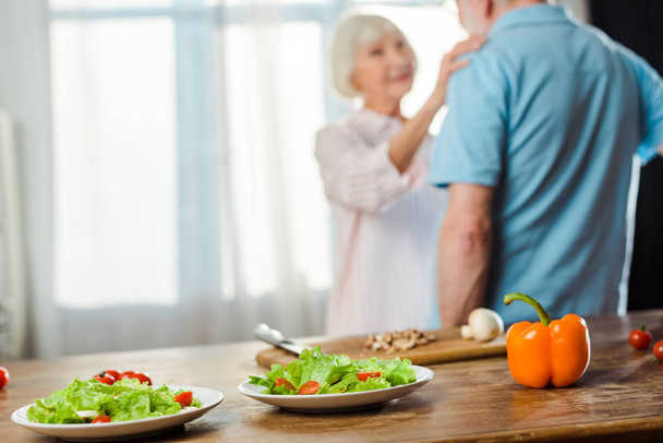 Selective focus of salad and fresh vegetables on kitchen table and senior couple at background - Photo, Image