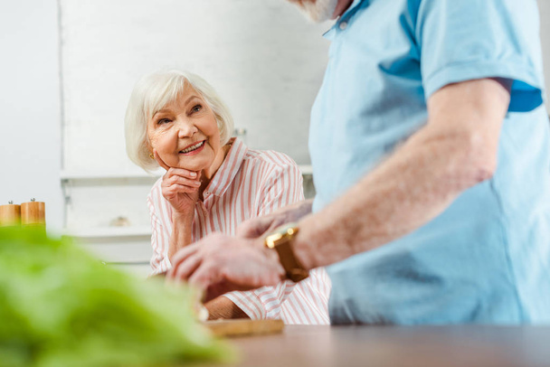 Szelektív fókusz mosolygó idősebb nő néz férj főzés közben a konyhaasztalon - Fotó, kép