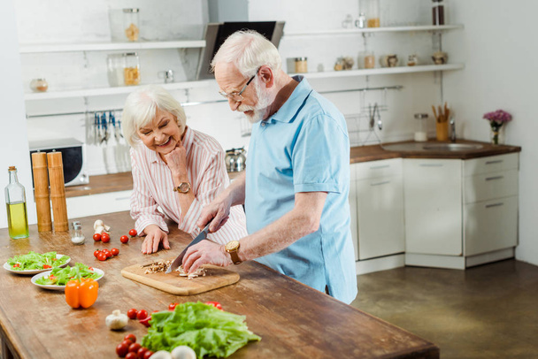 Sonriente mujer mayor de pie por el marido cortar verduras en la mesa de la cocina
 - Foto, Imagen