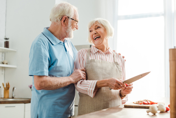 Senior man hugging smiling wife while cutting vegetables on kitchen table - Φωτογραφία, εικόνα