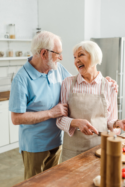 Επιλεκτική εστίαση του ηλικιωμένου άνδρα αγκαλιάζει χαμογελαστή γυναίκα κατά τη διάρκεια μαγείρεμα στο τραπέζι της κουζίνας - Φωτογραφία, εικόνα