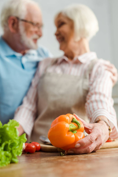 Selective focus of senior woman holding bell pepper while smiling at husband during cooking in kitchen - Zdjęcie, obraz