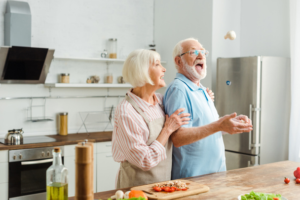 Senior couple laughing while man throwing up mushroom by fresh vegetables on kitchen table - Photo, Image